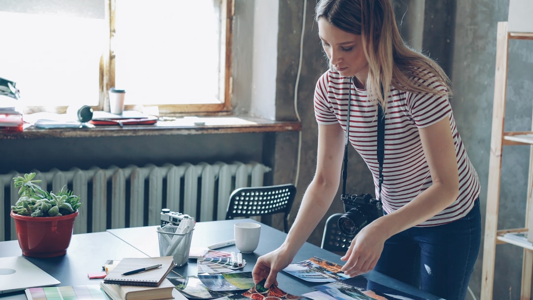 Young blond female photographer is making flatlay from colorful photos on table and shooting them with camera. She is displaying pictures near color palette and markers on desk.