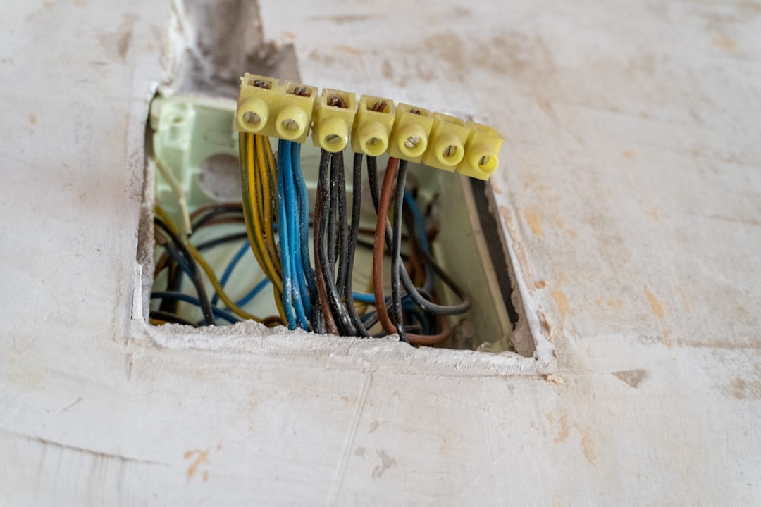 A close-up of an open electrical junction box set into a plaster wall, where neatly aligned colored wires are secured by a terminal block, revealing the hidden order behind interior wiring. The image conveys precision, safety, and the technical backbone of home renovation and electrical work.