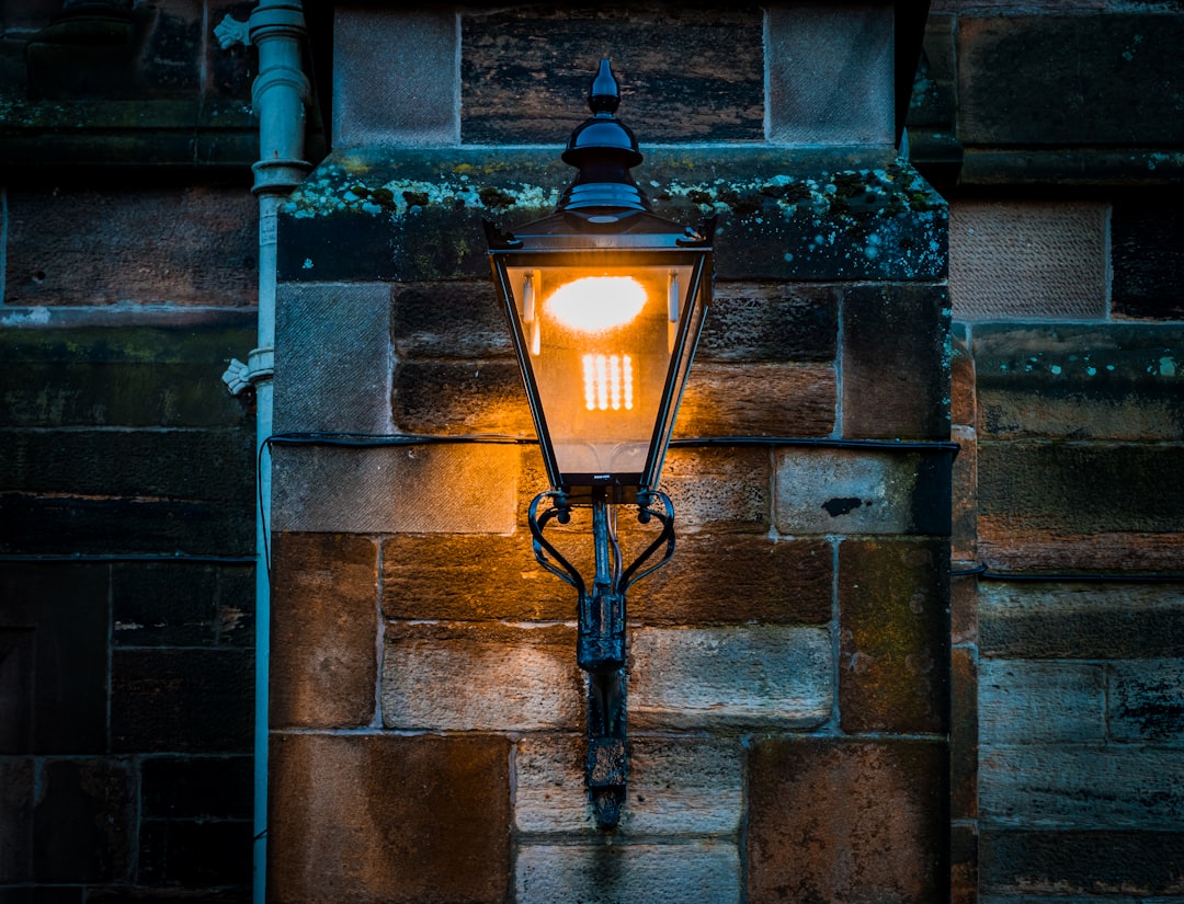 A vintage wall lamp affixed to the mottled stonework of the University of Glasgow, its light casting a soft glow that highlights the texture of the historic walls. This lamp is one of many that line the campus, contributing to the university's charming atmosphere.