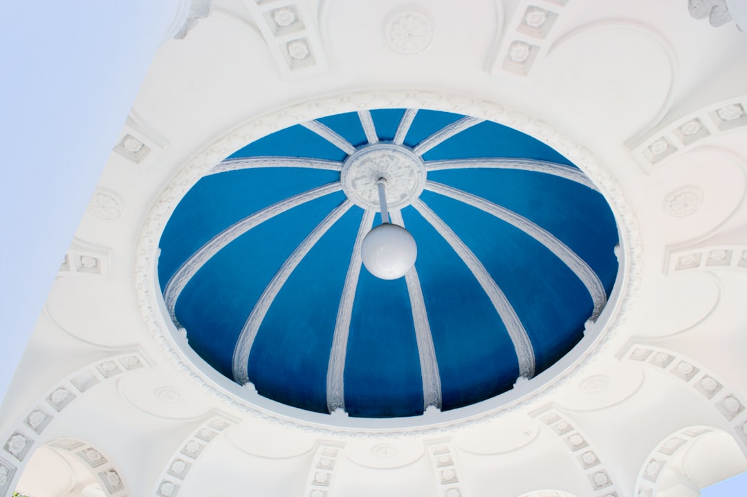 A striking upward view of an ornate architectural dome painted in vivid blue with white radial ribs, set within an elaborately decorated white stone frame. A spherical lamp hangs from the center, emphasizing the geometric precision and classical elegance.