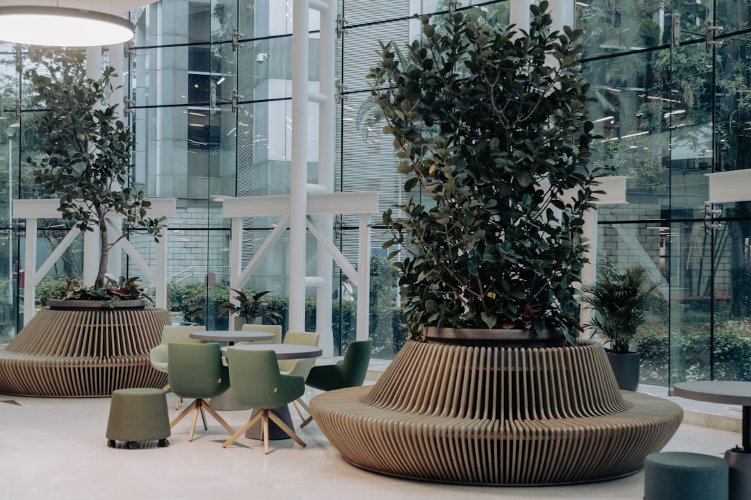 The image shows a modern office lobby or waiting area with a contemporary design. The space features large floor-to-ceiling windows that allow natural light to flood the area, creating a bright and airy atmosphere. The seating arrangement includes round tables surrounded by green chairs, and stylish planters with lush indoor plants. The planters have unique, ribbed designs that add an architectural element to the space. The decor is minimalistic, with a focus on functionality and comfort. The office lobby provides a welcoming environment, combining modern aesthetics with natural elements.