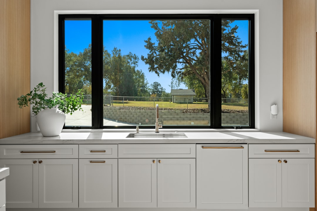 Modern kitchen sink with white shaker cabinetry, brass hardware, and large black-framed picture window overlooking greenery. Bright contemporary home interior featuring marble countertops, natural light, and clean minimalist design. High-end residential kitchen with timeless finishes and warm wood accents.